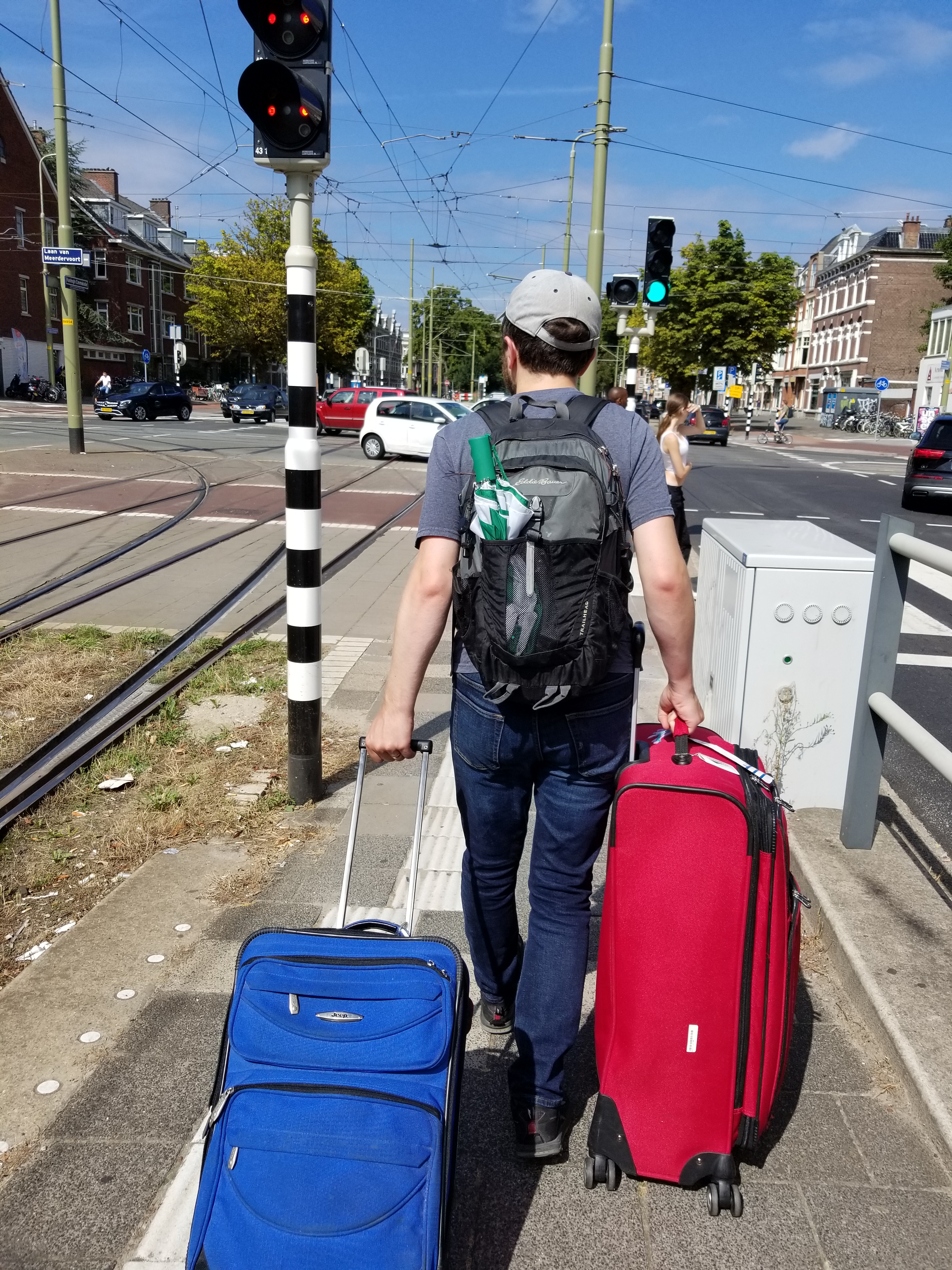 I walk away from the camera while pulling two large suitcases, one red and one blue. We are walking down the middle walkway of a busy Dutch street.