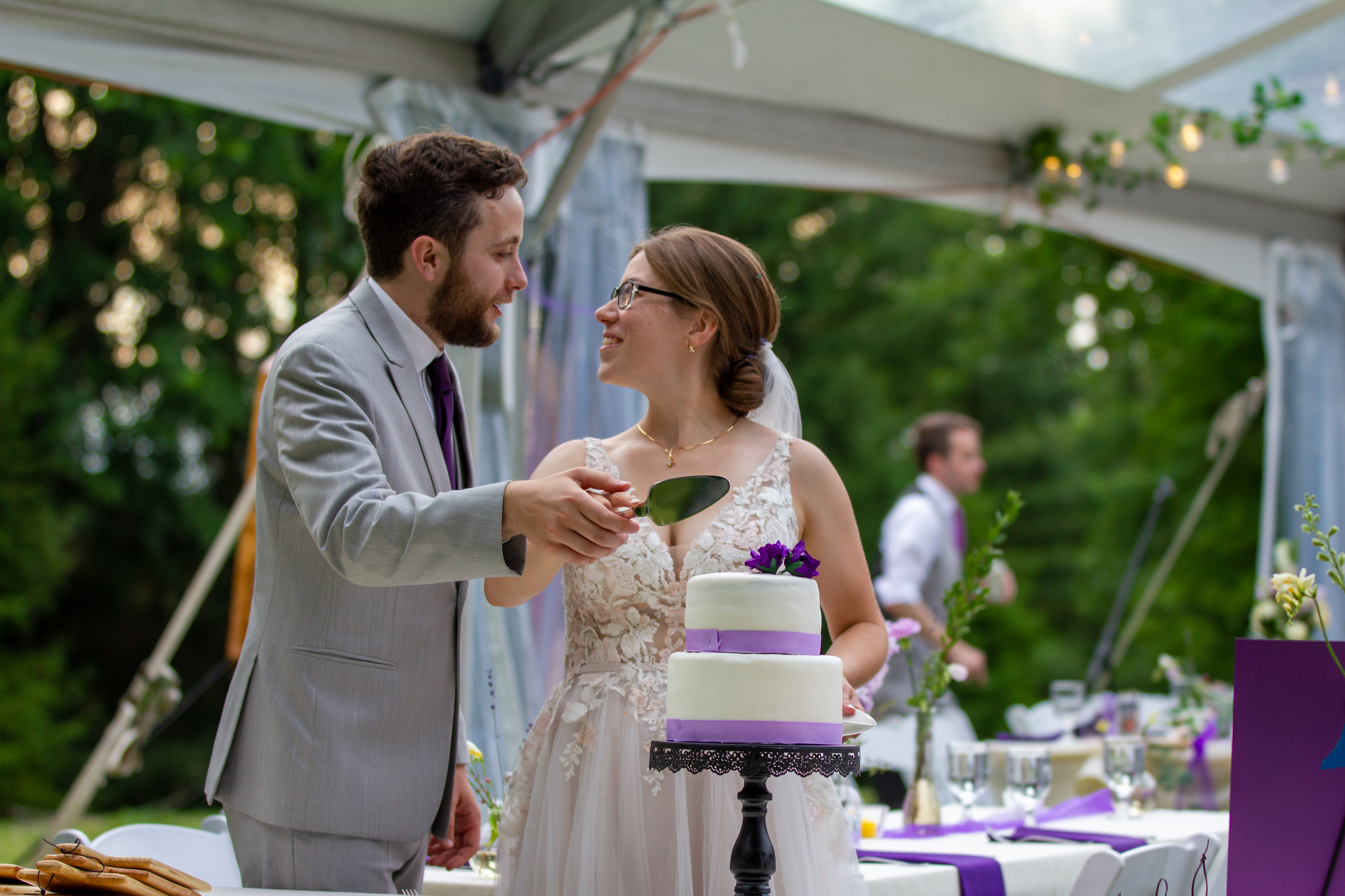 I hold my wife Julia's hand as we prepare to cut the cake for our wedding. She's in a white wedding gown and I'm in a grey suit. The white cake is adorned with purple flowers and ribbons.